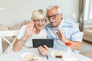 Happy older couple using computer tablet together at home, excited mature man and woman looking at mobile device screen, video call chatting online while having breakfast at home. photo