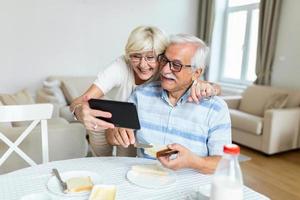 Senior couple smiling and looking at the same tablet. Old couple having video call with friends or family during breakfast photo