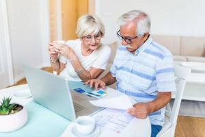 Happy middle aged husband and wife sitting at table with laptop and paper bills, calculating domestic incomes together at home. photo