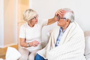 mujer comprobando la temperatura de la fiebre del hombre mayor. el viejo esposo descansa en casa sintiéndose enfermo mientras su esposa revisa la fiebre tocando la frente foto