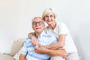 retrato amante de la esposa mayor abrazando al marido sentado en un cómodo sofá. feliz pareja madura sonriendo y mirando la cámara, posando para una foto familiar en casa. pareja de ancianos sintiéndose feliz