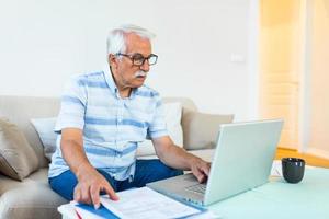 un anciano de cabello gris enfocado se sienta en el sofá leyendo notificaciones bancarias calculando los gastos domésticos, un hombre maduro moderno concentrado considera el papeleo financiero, paga facturas en una laptop en línea foto