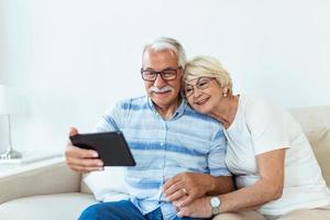 feliz pareja mayor usando tableta de computadora juntos en casa, emocionado hombre y mujer maduros mirando la pantalla del dispositivo móvil, comprando o chateando en línea, sentados en un acogedor sofá en la sala de estar foto