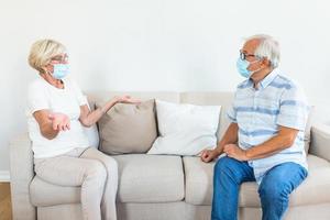 Two Elderly friends sitting in social distance wearing protective mask and talking on the sofa, preventing covid 19 coronavirus pandemic infection spread. Social distancing on sofa at home photo