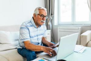 Old retiree man sit on couch holding receipt paper calculates on calculator monthly expenses, elderly male managing budget, planning finances, analyzes charges, check utility bills concept photo