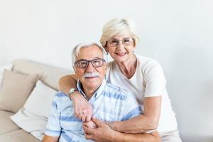 Portrait loving older wife hugging husband sitting on cozy couch. Happy senior mature couple smilling and looking at camera, posing for family photo at home. Elderly couple feeling happy