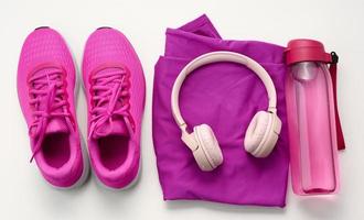 par de textil púrpura Deportes zapatillas, inalámbrico auriculares, un toalla y un botella de agua en un blanco antecedentes. ropa de deporte foto
