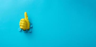 a hand in a yellow household rubber glove sticks out of a torn hole and shows a gesture okay, thumb up. Blue background photo