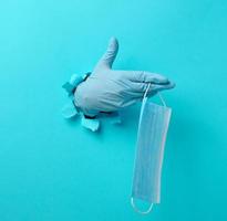 a female hand holds a disposable medical mask to protect against viruses during an epidemic and pandemic. Part of the body sticks out of a torn hole in blue paper photo