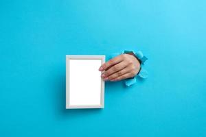 female hand holding empty white wooden frame, body part sticking out of torn hole in blue paper background photo