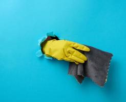 hand in a yellow rubber cleaning glove holds a dry rag on a blue background. Part of the body stick out of the hole photo