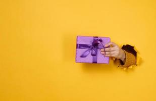 female hand holds a box with a gift on a yellow background, part of the body sticks out of a torn hole in a paper background. Congratulation, holiday surprise photo