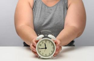plump woman sits at a table and holds a round white alarm clock. The concept of fractional nutrition, time to lose weight, adherence to sleep photo