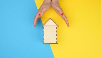 dos hembra manos doblada a cada otro terminado un de madera miniatura modelo casa en un amarillo azul antecedentes. real inmuebles seguro concepto foto