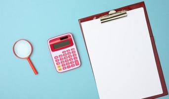 pink calculator, folder with blank white sheets and black magnifier on a blue background photo