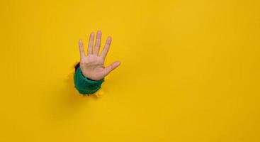 woman's hand with an open palm sticks out of a torn hole in yellow paper, gesturing to stop photo