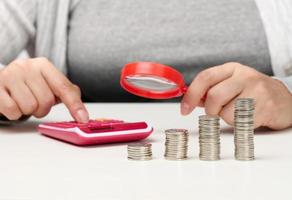 a woman sits at a table and counts on a calculator in the other hand a magnifying glass, a stack of coins on the table. Calculation of expenses and income photo
