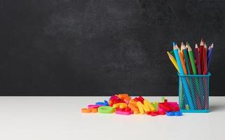 metal glass with pens, pencils and felt-tip pens and scissors on the background of an empty black chalk board, white table photo