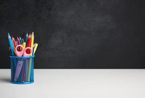 metal glass with pens, pencils and felt-tip pens and scissors on the background of an empty black chalk board, white table photo