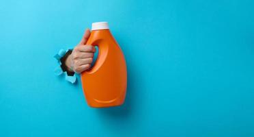 orange plastic bottle with liquid detergent in a female hand on a pink background. A part of the body sticks out of a torn hole in the background photo