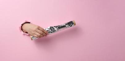 female hand holding a scroll of gray food foil on a pink background, part of the body sticking out of a torn hole photo