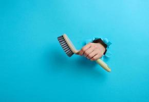 female hand holds a gray plastic brush on a blue background. A part of the body sticks out of a hole with torn edges in a paper background photo