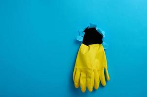 un par de amarillo látex casa limpieza guantes pega fuera de el Rasgado agujero de el azul papel antecedentes foto