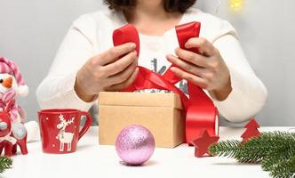 un mujer en un blanco suéter se sienta a el mesa y envuelve regalos para Navidad. preparando para el Días festivos foto