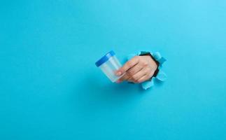 female hand holds an empty plastic jar for urine tests, the hand is sticking out of a torn hole in a blue background photo