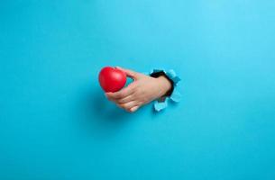 female hand holding a red heart on a blue background. Part of the body sticks out of the torn paper photo
