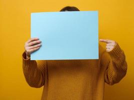 woman in an orange sweater holds a blank sheet of paper on a yellow background. Place for an inscription, advertisement, information photo