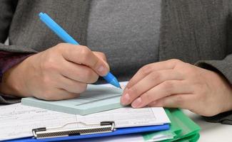 woman of Caucasian appearance sits in gray clothes at a white table and signs documents with a metal pen. Businessman at work, signature on contract and invoice photo