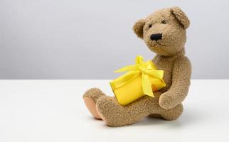 cute brown teddy bear holding a box wrapped in yellow paper and silk ribbon on white background. Prize and congratulations photo