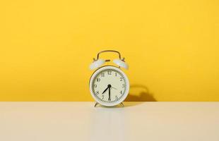 un redondo alarma reloj soportes en un blanco mesa, medio pasado Siete en el Mañana. amarillo antecedentes foto
