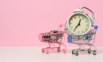 round alarm clock in a miniature shopping cart with change on a white table. Concept time is money, waste of money and poverty photo