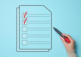 a female hand holds a red market and a document with cells for marking on a blue background. Voting concept, data validation, shopping list photo