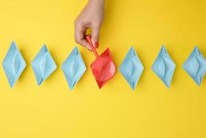 female hand holds a magnifying glass over a row of paper boats on a yellow background. Talent search concept photo