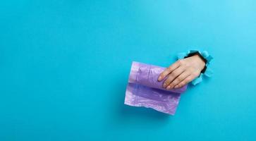 female hand holds a bundle of transparent garbage bags on a blue background. A part of the body sticks out of a torn hole in the paper photo