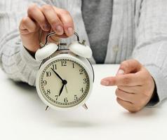 female hand holds a white metal alarm clock on a white table, time five minutes to seven in the morning photo