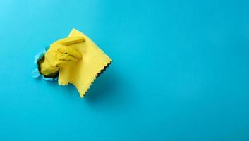 a hand in a yellow rubber cleaning glove holds a dry rag on a blue background. Part of the body stick out of the hole in the paper, banner photo