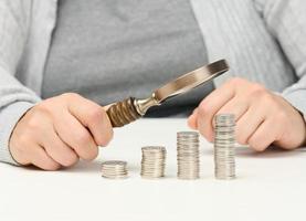 a woman sits at a table and holds a wooden magnifying glass over stacks of white coins. The concept of budget planning, savings. Investment search photo