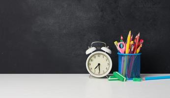 white round alarm clock and a metal glass with pens, pencils and felt-tip pens on the background of an empty black chalk board photo