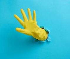mano en un amarillo látex limpieza guante palos fuera de un Rasgado agujero en un azul papel antecedentes foto