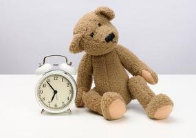 brown teddy bear sits on a white table and a round alarm clock, at five minutes to seven in the morning photo