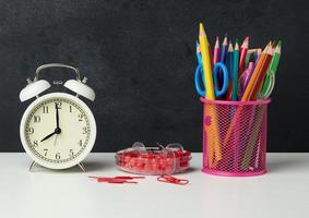 white round alarm clock and a metal glass with pens, pencils and felt-tip pens on the background of an empty black chalk board photo