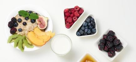 leche, plato con gachas de avena y fruta, jugo recién exprimido en un decantador de vidrio transparente, miel en un bol sobre una mesa blanca. desayuno saludable foto