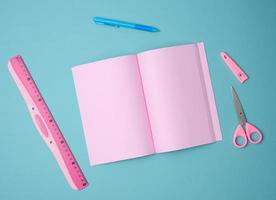 open notebook with blank pink sheets, scissors and ruler on blue background, top view photo