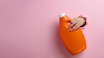 naranja el plastico botella con líquido detergente en un hembra mano en un rosado antecedentes. un parte de el cuerpo palos fuera de un Rasgado agujero en el fondo, un sitio para un inscripción foto