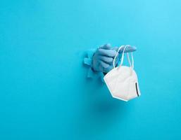 female hand holds a white disposable medical mask to protect against viruses during an epidemic and pandemic. Part of the body sticks out of a torn hole in blue paper photo