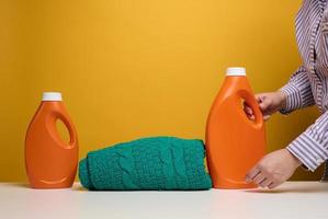 ropa doblada lavada y botellas grandes de plástico naranja con detergente líquido sobre una mesa blanca, fondo amarillo. tarea de rutina foto